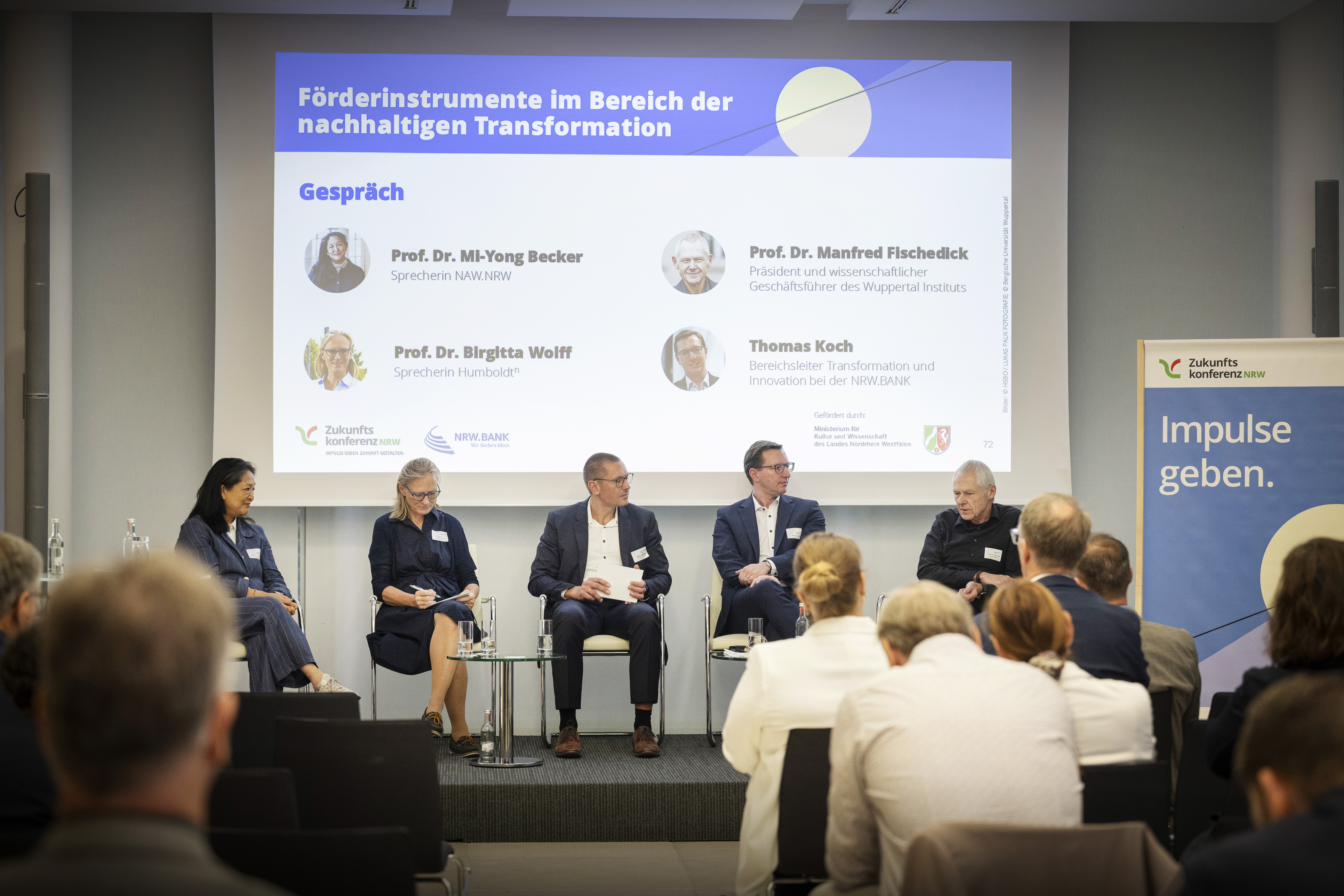 Fünf Personen sitzen auf einer Bühne und führen ein Gespräch, hinter ihnen wird eine Präsenattion an die Wand projeziert und vor ihnen sitzen mehrere Menschen im Publikum und hören ihnen zu.