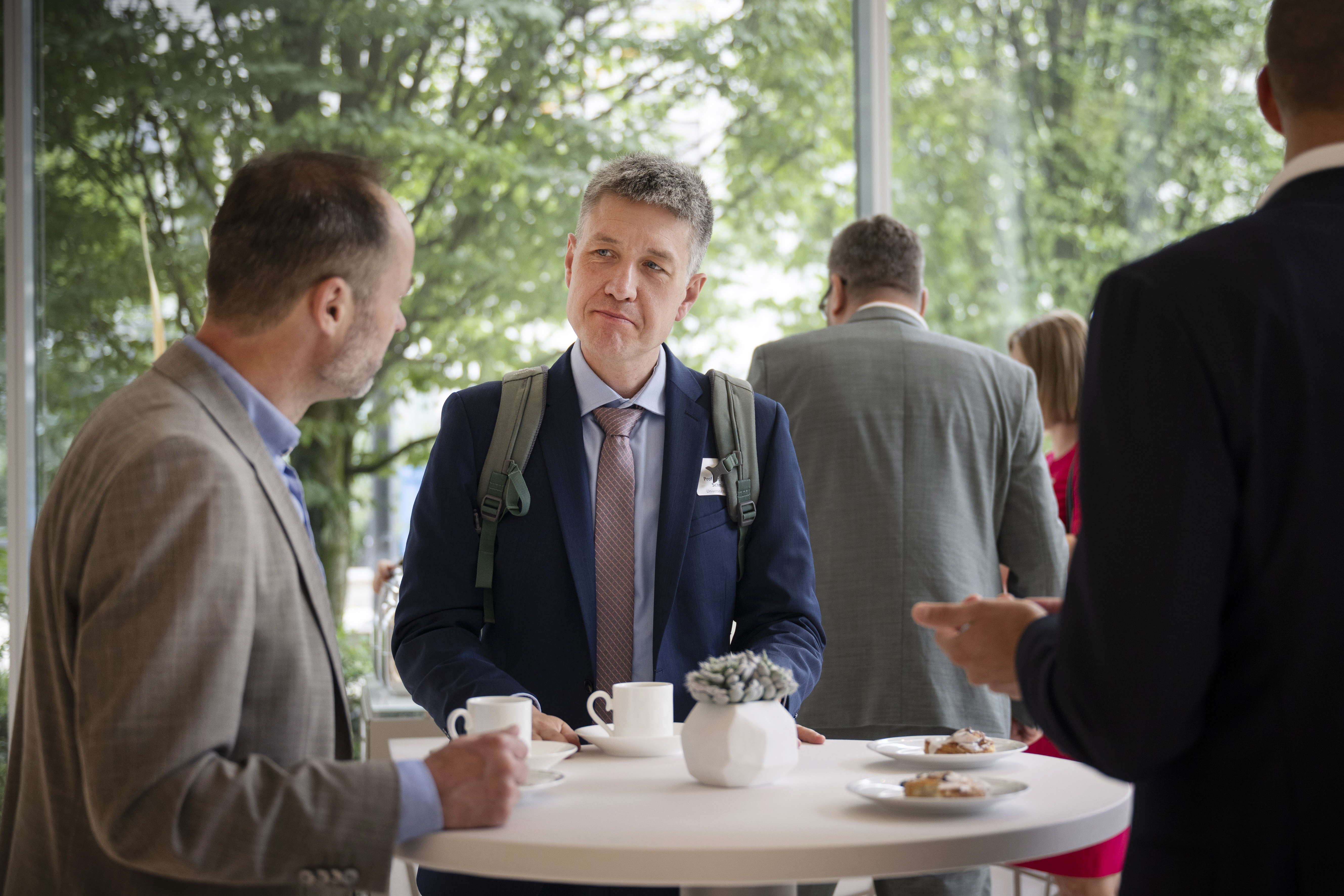 Zwei Männer stehen an einem Stehtisch. Sie tragen beide einen Anzug und trinken Kaffee. Der linke Mann spricht, während der rechte interessiert zuhört.