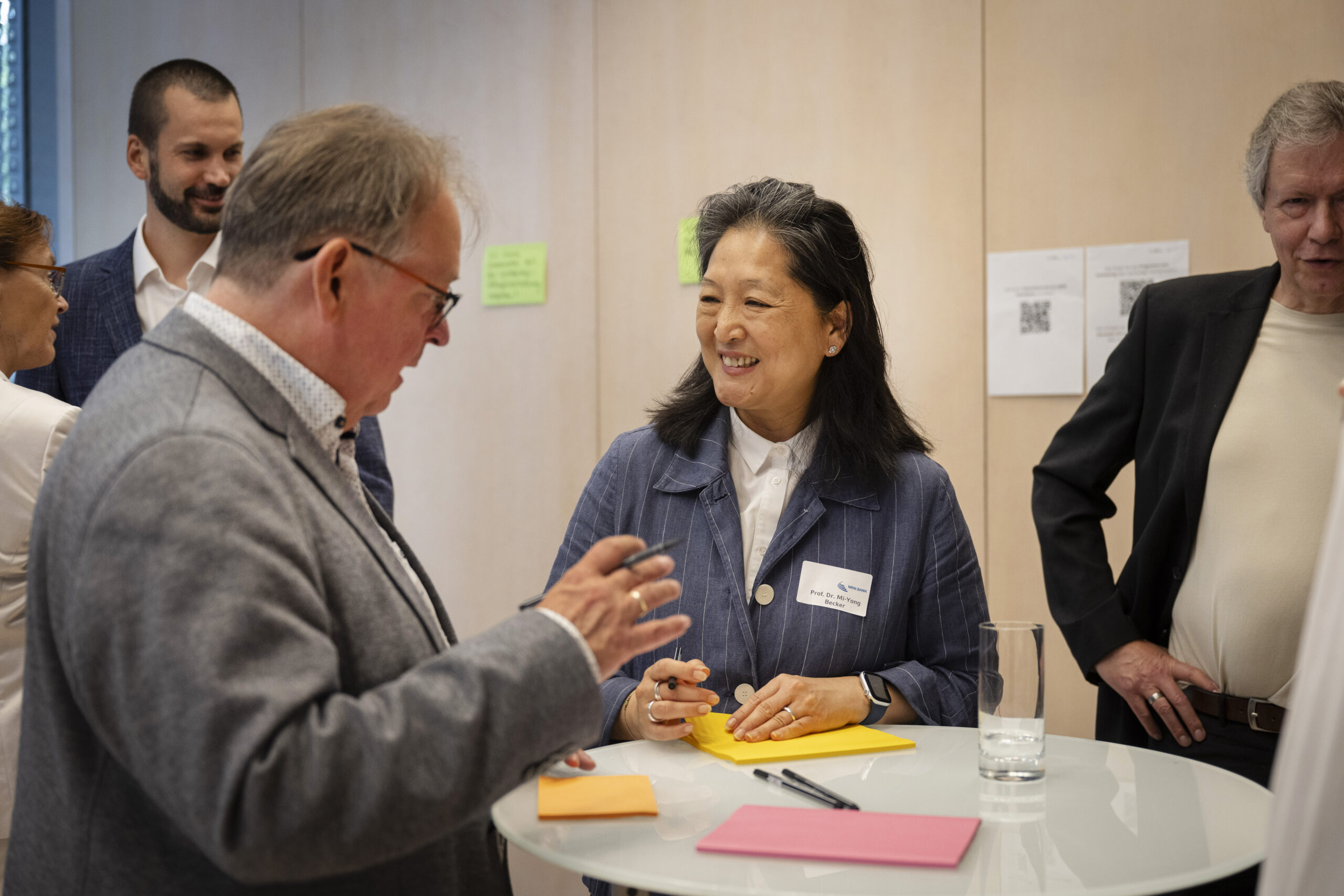 Eine Frau und ein Mann stehen an einem Stehtisch, unterhalten sich lächelnd und beschriften Post-Its die vor ihnen liegen. Im Hintergrund stehen weitere sich unterhaltende Menschen.
