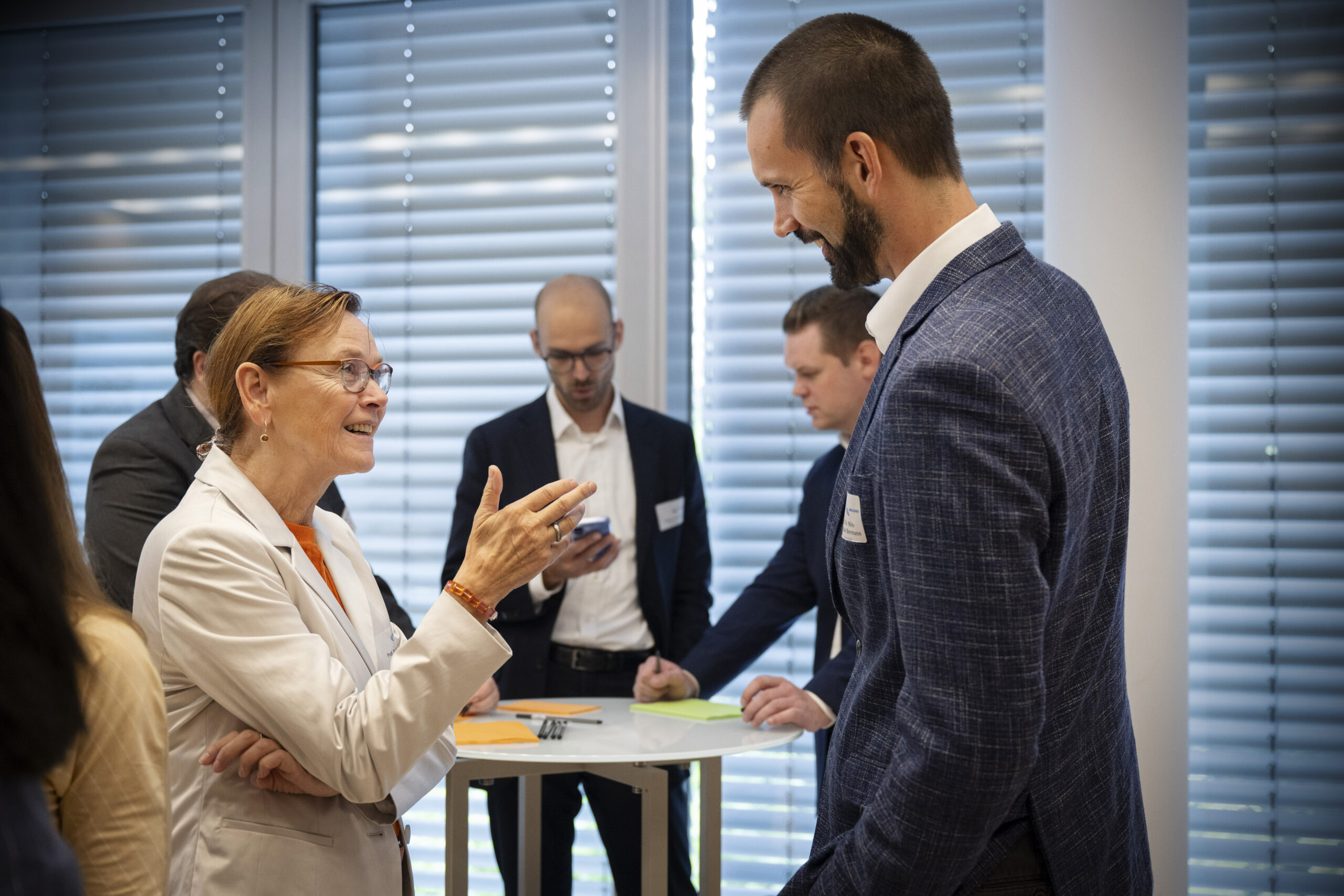 Eine Frau und ein Mann unterhalten sich lächelnd. Beide tragen ein Jacket. Im Hintergrund stehen drei weitere Männer um einen Stehtisch herum und beschriften vor ihnen liegende Post-Its.