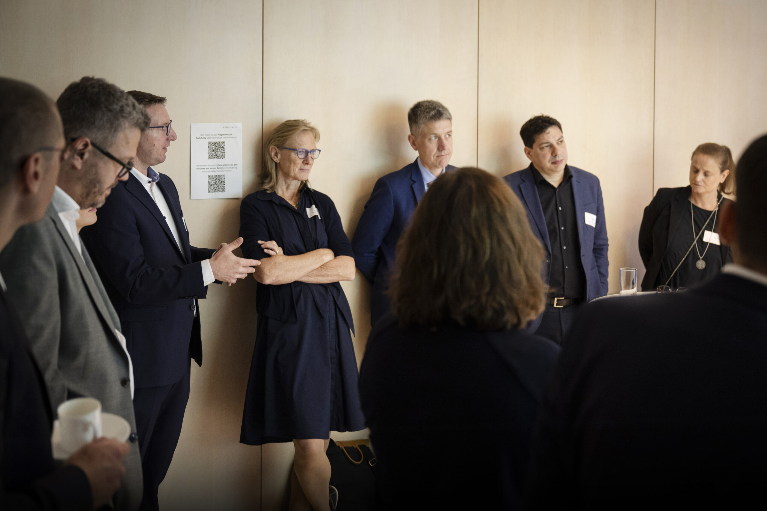 Eine große Gruppe an Menschen steht in einem Raum und diskutiert aufmerksam. Alle Personen tragen elegante Kleidung.