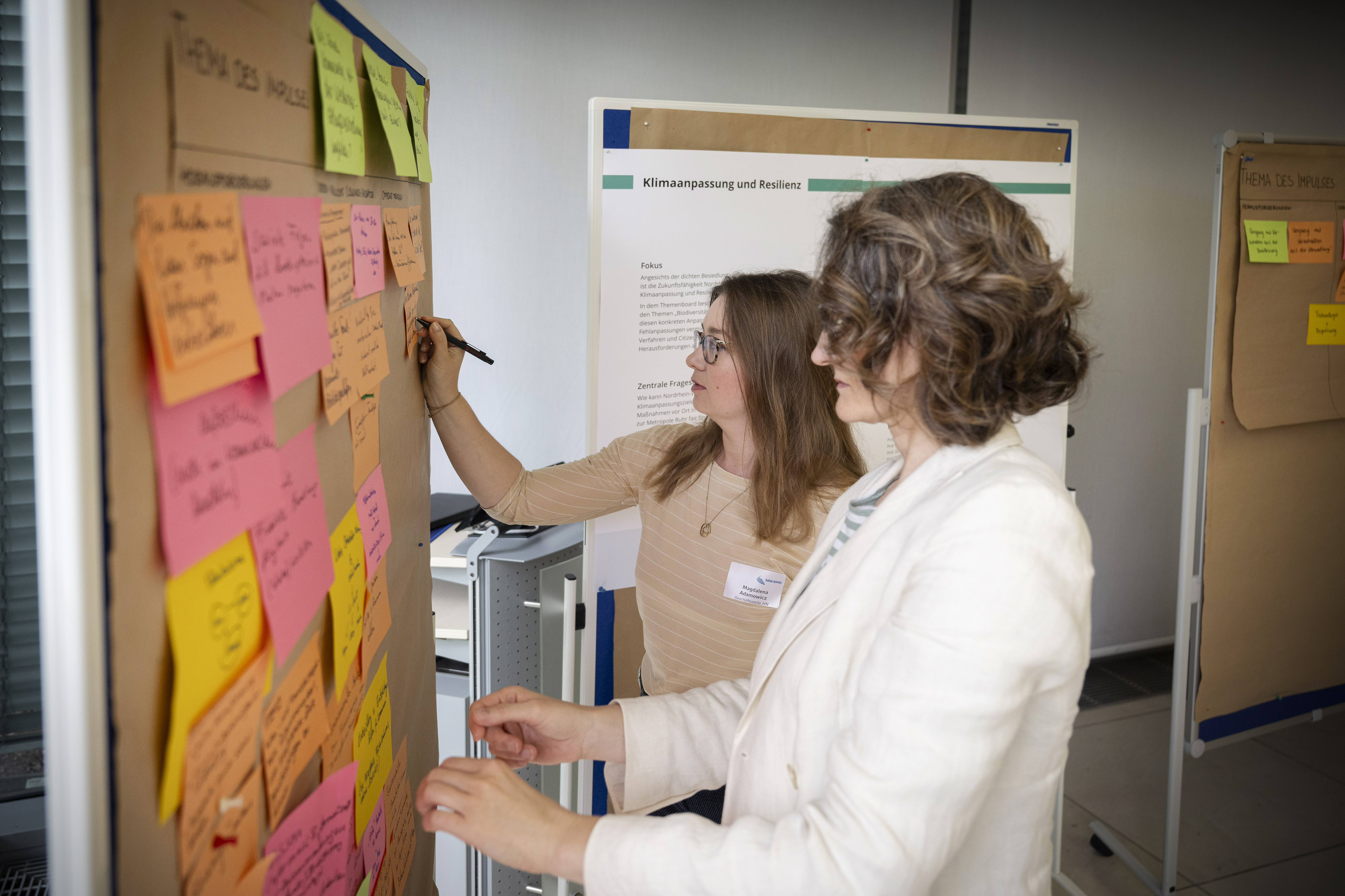 Zwei Frauen stehen vor einer Metaplanwand, an der ein Poster mit vielen bunten Post-Its hängt und arbeiten an den Materialien. Im Hintergrund stehen weitere Metaplanwände mit Postern.
