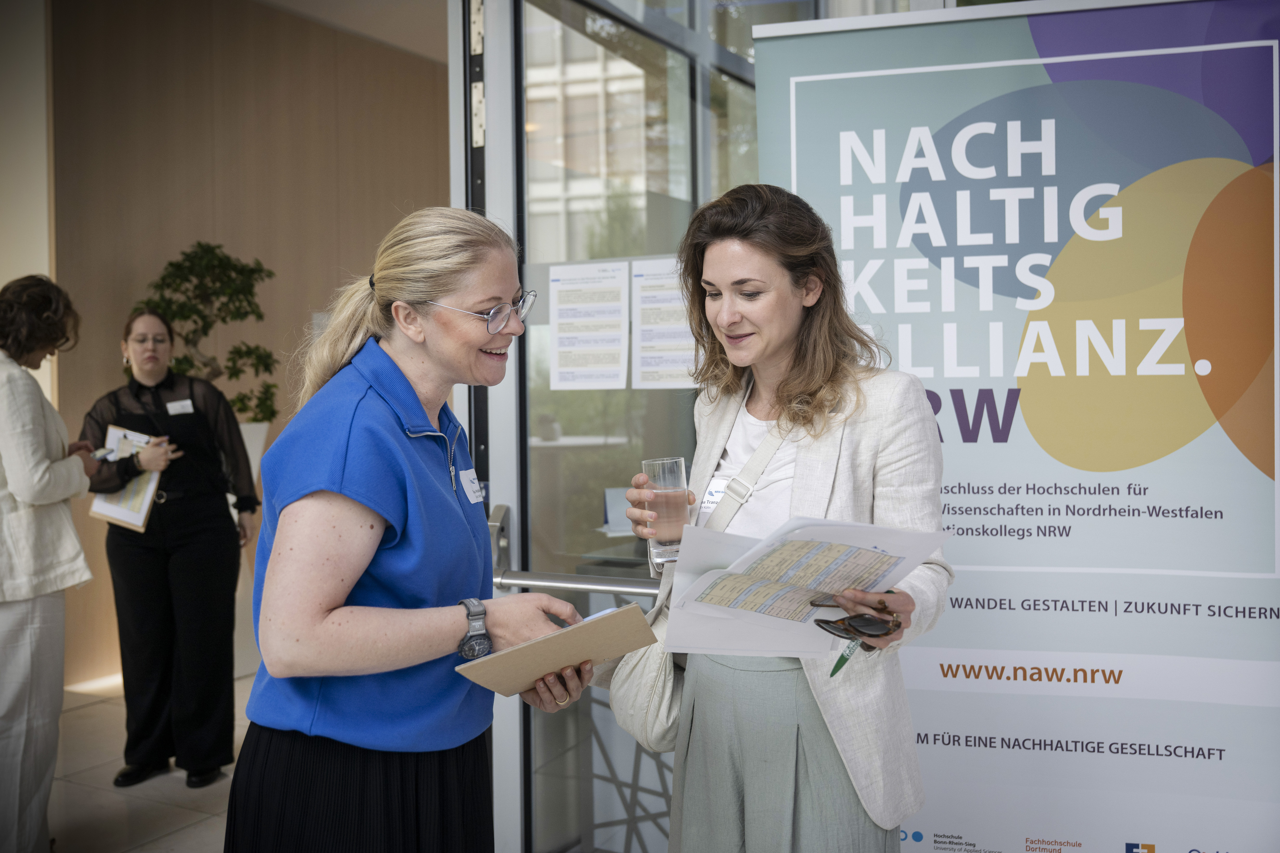Zwei Frauen stehen vor einem Roll-Up der Nachhaltigkeitsallianz NRW und blicken auf einen Zettel den die eine der beiden Frauen in der Hand hält. Beide lächeln.