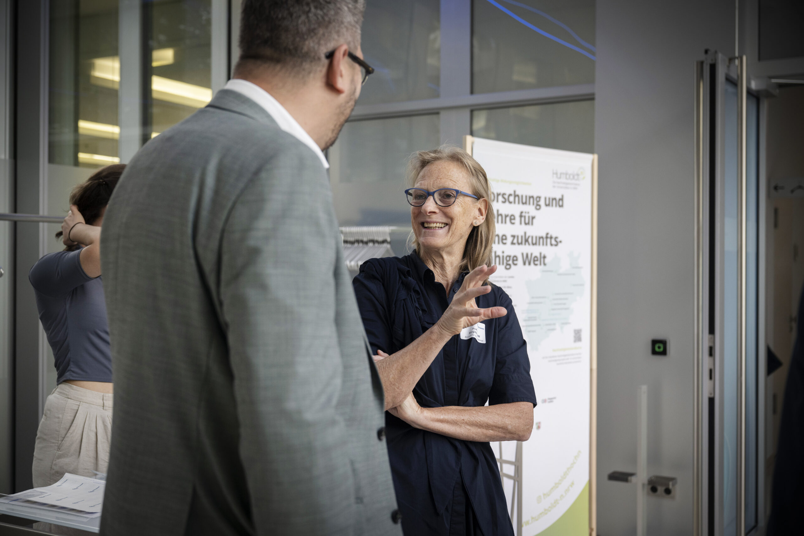 Eine blonde Frau mir blauer Brille und einem dunkelblauen Kleid gestikuliert lachend, während sie sich mit einem Mann in grauem Sacko unterhält, der mit dem Rücken zur Kamera steht.