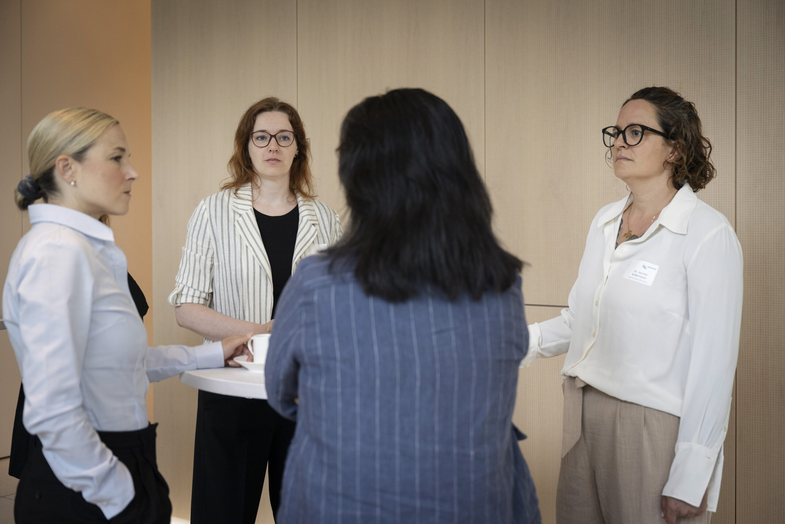 Vier Frauen stehen um einen Stehtisch herum und hören einer der vier Frauen, die mit dem Rücken zur Kamera steht, interessiert zu.