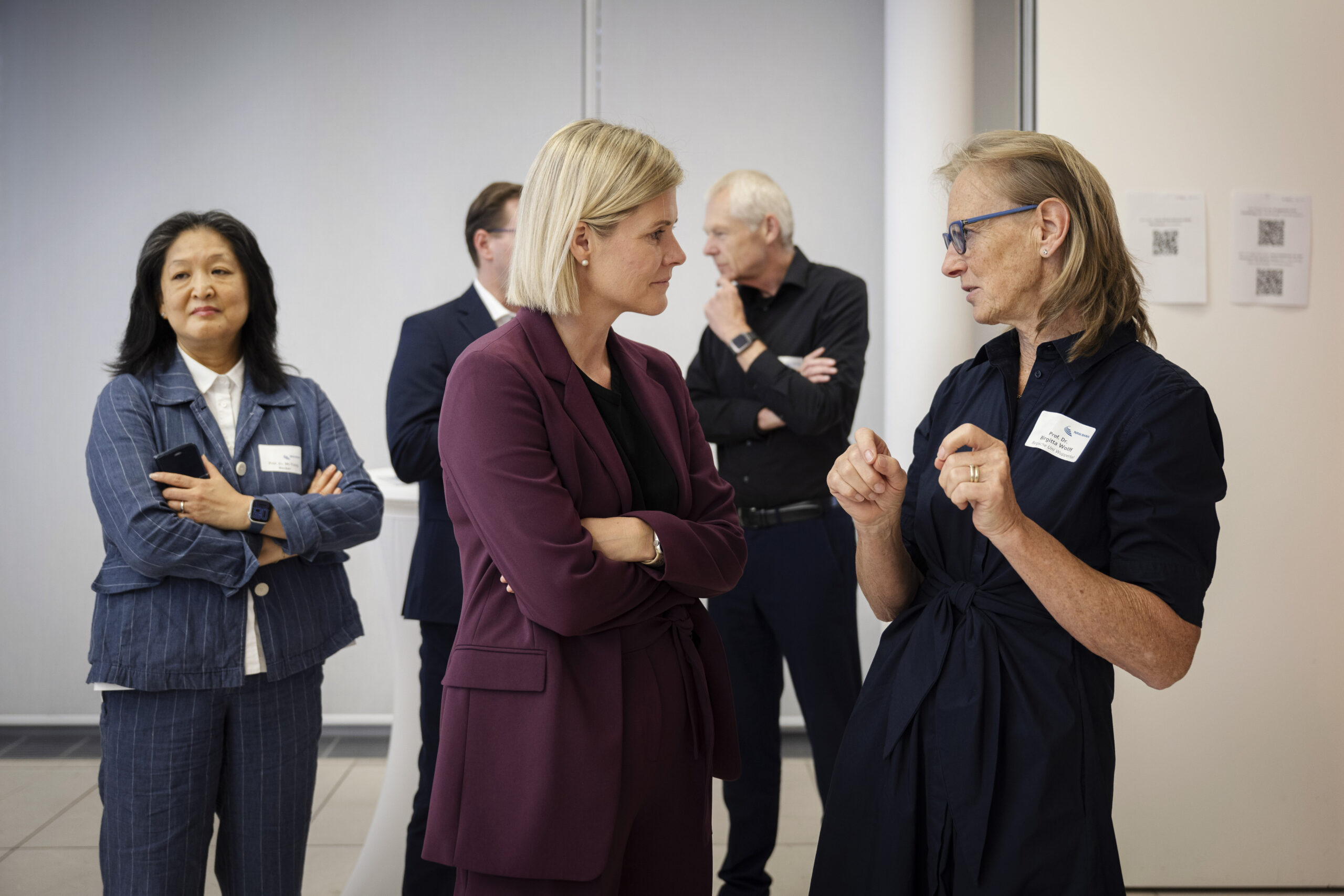 Zwei Frauen unterhalten sich angeregt. Die linke Frau trägt einen dunkelroten Blazer und blonde, kinnlange Haare, die rechte Frau trägt ein dunkelblaues Kleid, eine blaue Brille und schulterlanges dunkelblondes Haar.