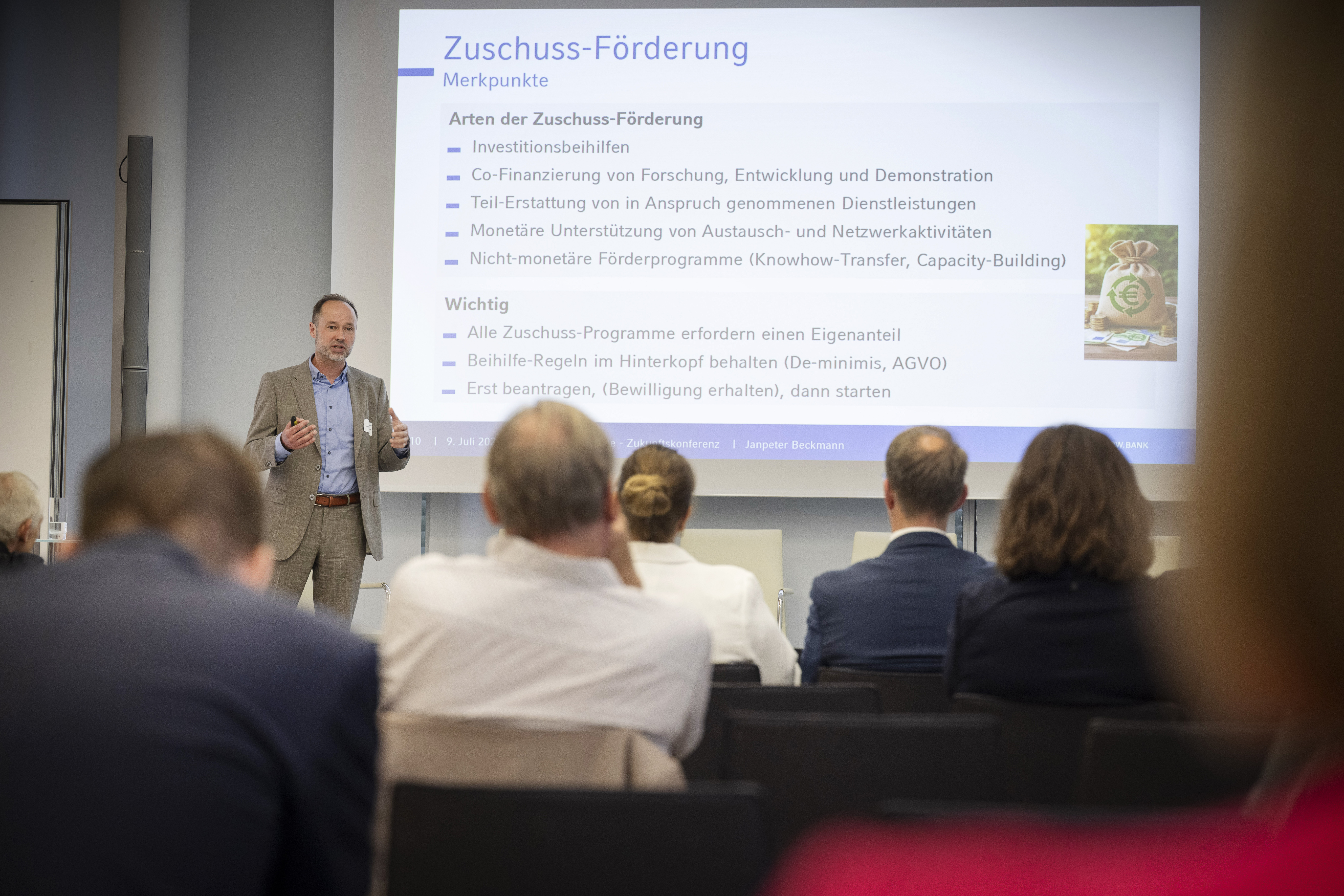 Ein Mann mit hellblauem Hemd und braun-grauem Sakko steht auf einer Bühne und hält eine Präsentation zum Thema "Zuschuss-Förderung". Hinter ihm werden die Folien an die Wand projeziert. Im Vordergrund sind die Hinterköpfe von einigen Menschen zu sehen, die dem Sprecher zuhören.