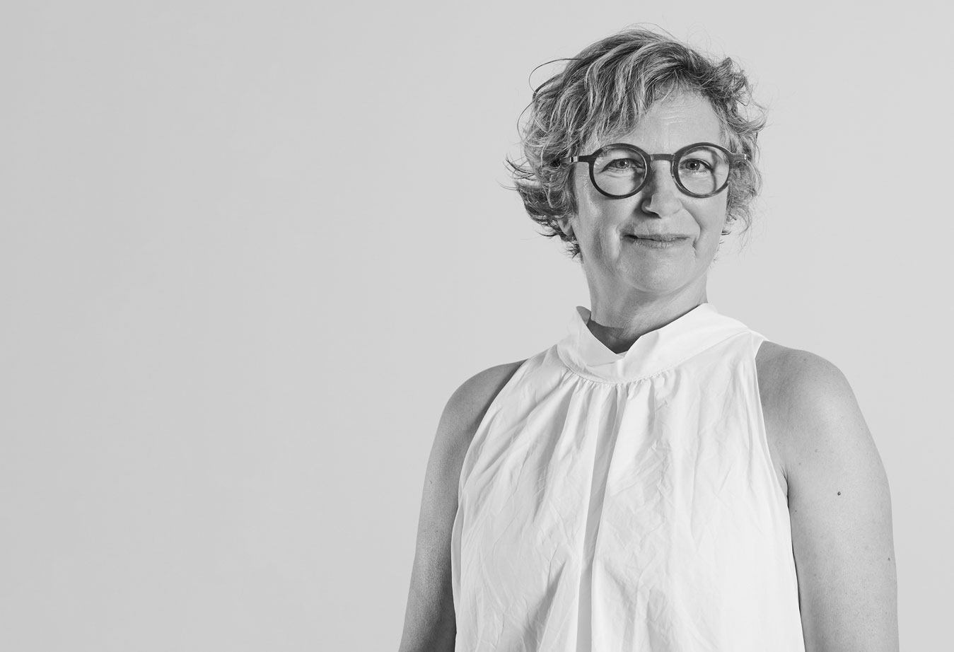 © www.heikefischer-fotografie.de Ein schwarz-weiß Foto einer Frau mit runder, dunkler Brille, grauen Locken und einer weißen Bluse, die in die Kamera lächelt.